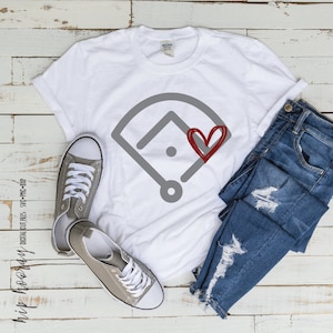 May include: White t-shirt featuring a grey baseball field graphic with a red heart on the front. The shirt is laid out on a white wooden surface with a pair of grey sneakers and blue jeans.