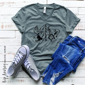 May include: Gray V-neck t-shirt with a black design of an eagle with a musical note and the word "EAGLES" printed vertically.