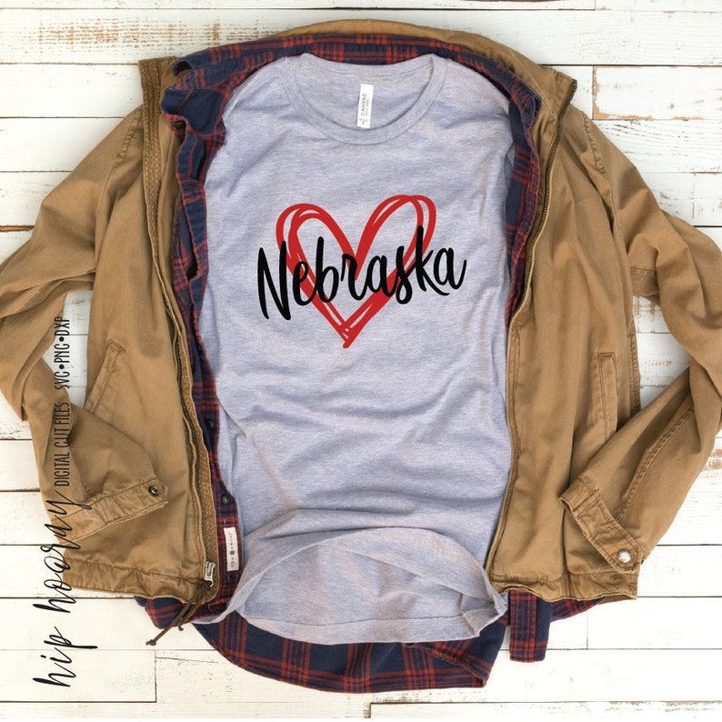 Peut inclure: Un t-shirt gris clair avec le mot "Nebraska" en &eacute;criture noire &agrave; l'int&eacute;rieur d'un c&oelig;ur rouge. La chemise est superpos&eacute;e &agrave; une veste marron et une chemise &agrave; carreaux rouge et bleu.