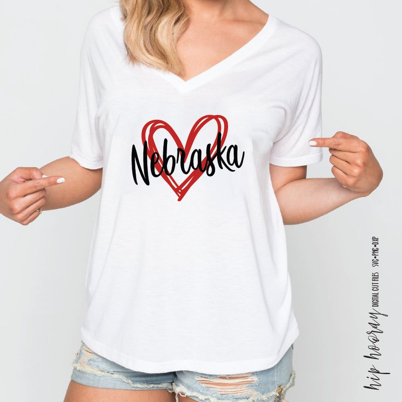 Peut inclure: T-shirt blanc &agrave; col en V avec un c&oelig;ur rouge et le mot "Nebraska" en &eacute;criture noire. Le design est de style simple et dessin&eacute; &agrave; la main. Un v&ecirc;tement d&eacute;contract&eacute; pour tous les jours.