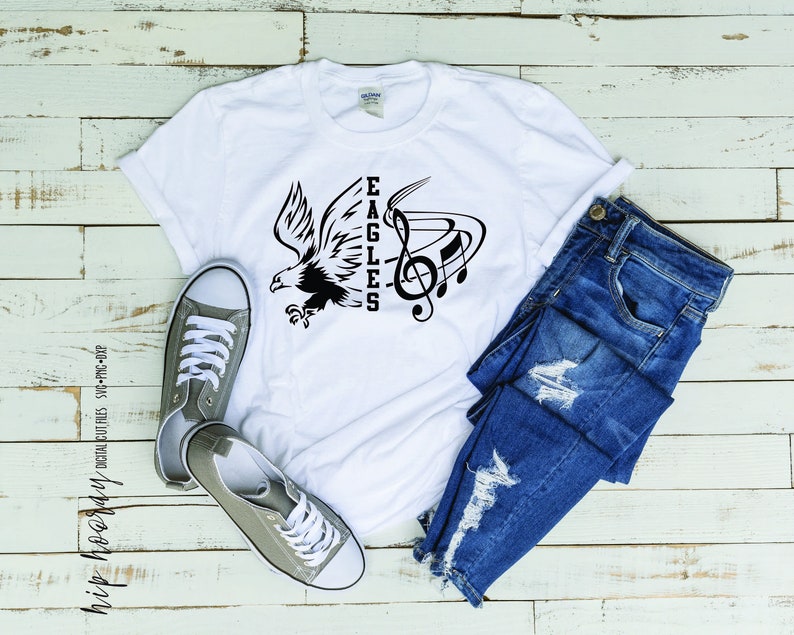 May include: White t-shirt with a black graphic of an eagle and musical notes with the word "EAGLES" written vertically. The shirt is laying on a wooden surface with a pair of grey canvas shoes and blue jeans.