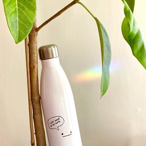 May include: A white insulated water bottle with a silver cap. The bottle has a small sticker with a speech bubble that says "I just want to be held." A small cartoon face is drawn below the speech bubble. The bottle is next to a plant.