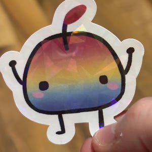 May include: A holographic sticker featuring a cartoon apple character with a rainbow gradient design. The apple has a black outline, small arms and legs, and rosy cheeks. A red stem and leaf are at the top.