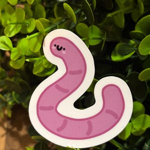 May include: A sticker depicting a cartoon worm in pink and white. The worm has a smiling face and segmented body. The sticker is set against a backdrop of green leaves.