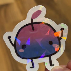 Könnte beinhalten: Ein holografischer Aufkleber mit einer Cartoon-Fruchtfigur mit einem Farbverlauf aus Rosa und Blau. Der Aufkleber hat einen schwarzen Umriss, kleine schwarze Augen und winzige Arme und Beine. Der Aufkleber wird von den Fingern einer Person gehalten.