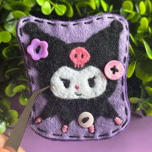 May include: A handmade felt character in a purple, black, and white color scheme. The character has a skull on its forehead and is decorated with pink and purple buttons. The edges are stitched with black thread.