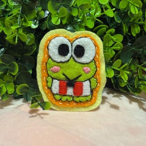 Könnte beinhalten: Ein handgefertigter Frosch-Anstecker aus Filz mit gelbem Rand und orangefarbenen Nähten. Der Frosch hat große weiße Augen, einen grünen Körper, rosa Wangen und einen rot-weiß gestreiften Mund. Vor dem Hintergrund von grünem Laub.