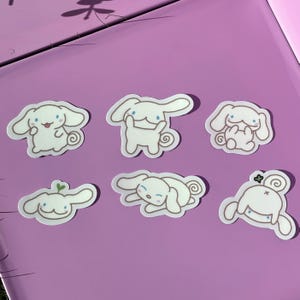 May include: Six white die-cut stickers featuring a cartoon character with long ears and a curly tail. The stickers are in various poses, including sitting, standing, and lying down. The stickers are on a purple surface.