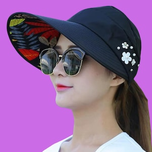 May include: Black sun hat with a wide brim featuring a colorful floral pattern on the underside. The hat is adorned with white flower and pearl embellishments. The hat is designed to provide sun protection.