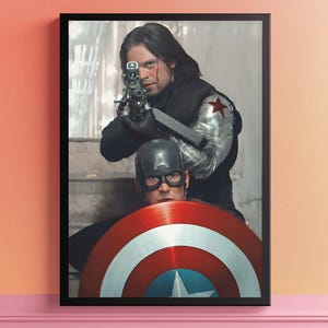 May include: Framed print featuring two characters from a superhero movie. One character aims a gun, while the other holds a shield with red, white, and blue stripes and a central star. The print is in a black frame.
