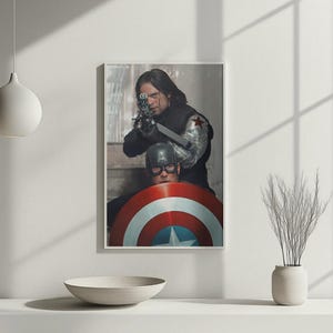 May include: Framed print featuring two figures from a superhero movie. One figure aims a gun, while the other is behind a shield with red, white, and blue stripes and a star. The print is displayed on a white surface.