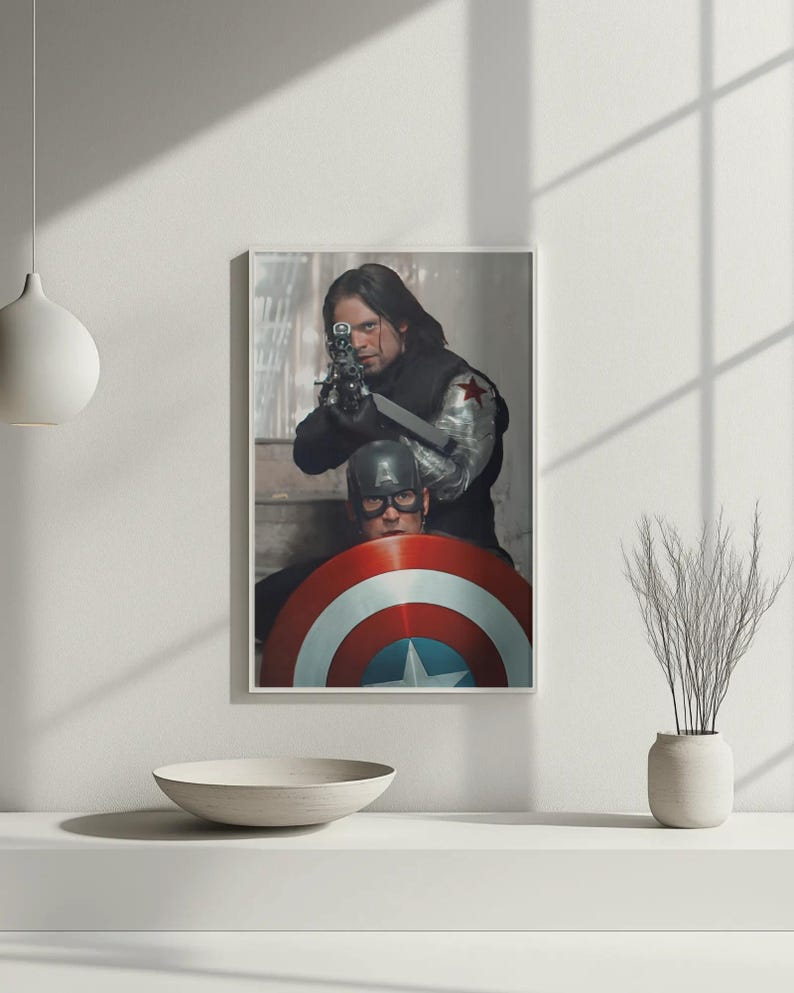 May include: Framed print featuring two figures from a superhero movie. One figure aims a gun, while the other is behind a shield with red, white, and blue stripes and a star. The print is displayed on a white surface.