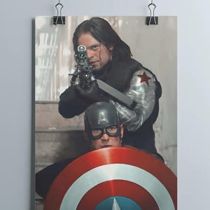 May include: A poster featuring two characters from a movie. One character is aiming a gun, wearing a black outfit with a metal arm and a red star. The other character is wearing a helmet and holding a shield with red, white, and blue stripes.
