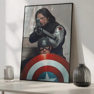 May include: Framed print featuring two characters from a superhero movie. One character is aiming a gun, while the other is holding a shield with red, white, and blue stripes and a star. The print is displayed on a white surface.
