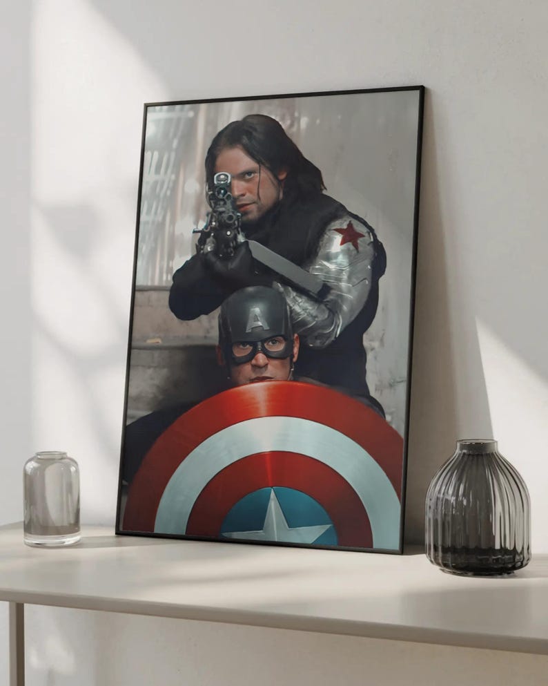 May include: Framed print featuring two characters from a superhero movie. One character is aiming a gun, while the other is holding a shield with red, white, and blue stripes and a star. The print is displayed on a white surface.