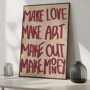 May include: A framed art print with the words "MAKE LOVE MAKE ART MAKE OUT MAKE MONEY" in red on a tan background. The print is displayed on a white shelf with a clear glass vase and a dark grey vase.