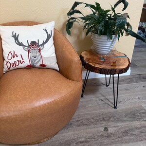 May include: A tan leather swivel chair with a square white throw pillow featuring a gray deer wearing a red and white patterned hat and the text Oh Deer! next to a round live-edge wood side table with black metal legs and a green plant.