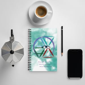 Peut inclure: Un carnet à spirales avec un fond turquoise aquarelle et un motif géométrique d'œil en violet, vert et rouge. Le texte "What you think, you become. What you feel, you attract. What you imagine, you create" est imprimé en dessous. Une tasse de café, un crayon, un téléphone et une cafetière italienne sont également présents.