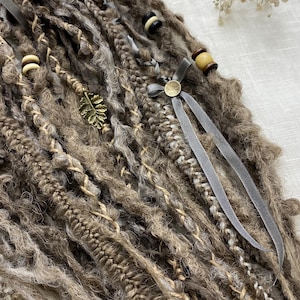 May include: A collection of brown and gray dreadlocks, adorned with decorative elements. The dreadlocks feature various textures and are accented with wooden beads, a gold-colored leaf charm, and gray ribbons. The overall aesthetic is natural and bohemian.