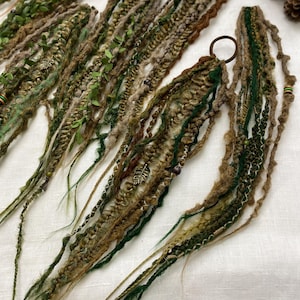 May include: Close-up of several handmade dreadlock extensions in shades of green, brown, and tan. The dreads are adorned with small green leaves, beads, and metal charms. Each dreadlock is secured with a brown hair tie. The overall aesthetic is natural and bohemian.