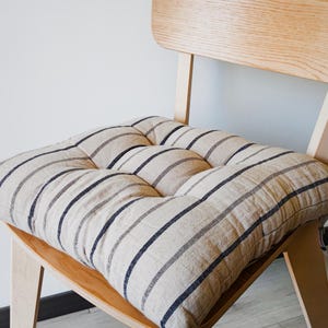 May include: A square seat cushion featuring a natural beige fabric with vertical navy blue stripes. The cushion is tufted with four circular indentations and is placed on a light-coloured wooden chair with a curved backrest and angled legs.
