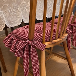 Puede incluir: Una silla de madera con respaldo de barrotes y un cojín de asiento capitoné con estampado de cuadros vichy en rojo y blanco. El cojín incluye un borde con volantes y lazos de tela que se sujetan al marco de la silla con nudos laterales.