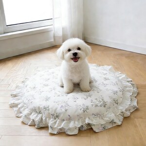 May include: A round pet bed featuring a cream-colored fabric with a delicate floral pattern in shades of green and purple. The bed is finished with a ruffled edge and a white lace trim, providing a soft and cushioned surface for a small animal.
