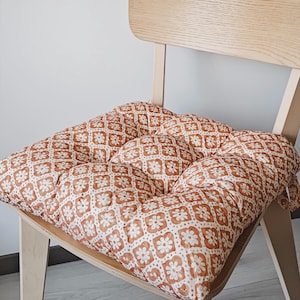 May include: A square chair cushion featuring a repeating pattern of white flowers on a terracotta orange background. The cushion is tufted with nine distinct sections and is placed on a light-colored wooden chair with a curved backrest.
