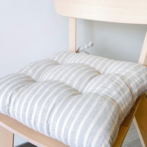 May include: A square chair cushion featuring a white and grey striped pattern. The cushion is tufted with visible stitching creating a grid of soft, padded sections. It includes fabric ties at the back corners for securing it to a wooden chair.