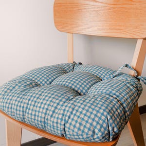 Può includere: Un cuscino per sedia quadrato e trapuntato con un motivo a quadretti vichy blu e bianchi. Il cuscino è fissato a una sedia in legno chiaro tramite lacci in tessuto sugli angoli posteriori, offrendo una seduta morbida con cuciture visibili.