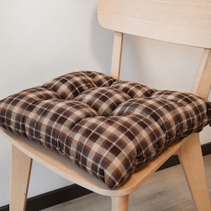 Puede incluir: Un cojín de silla cuadrado con un patrón de cuadros en tonos marrón, beige y blanco. El cojín está capitoné con nueve hendiduras circulares distintas en su superficie, creando una apariencia acolchada y segmentada sobre una silla de madera clara.