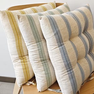 Könnte beinhalten: Drei quadratische Sitzkissen mit vertikalen Streifen in Gelb, Grün und Blau auf hellem Beige. Die Kissen liegen auf einem Holzstuhl und zeigen ein schlichtes, neutrales Design.