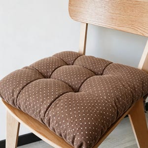 Könnte beinhalten: Ein quadratisches braunes Sitzkissen mit weißem Punktemuster, das ein gestepptes Design mit neun verschiedenen Abschnitten aufweist. Das Kissen liegt auf einem hellen Holzstuhl mit einer geschwungenen Rückenlehne und vier stabilen Beinen.