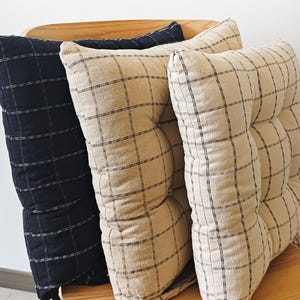 Könnte beinhalten: Drei quadratische Sitzkissen mit Karomuster. Ein Kissen ist schwarz mit weißen Gitterlinien, während die anderen beiden beige mit schwarzen Gitterlinien sind. Jedes Kissen hat ein gestepptes Design mit sichtbaren Nähten in einem Quadratmuster.
