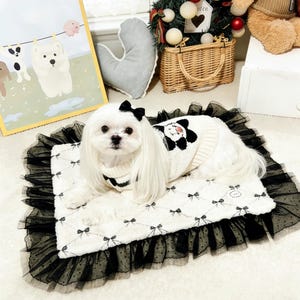 May include: A small white dog wearing a sweater with a panda design and a black bow. The dog is lying on a white and black bow-patterned pet bed with a black lace trim. A framed picture of dogs hangs in the background.