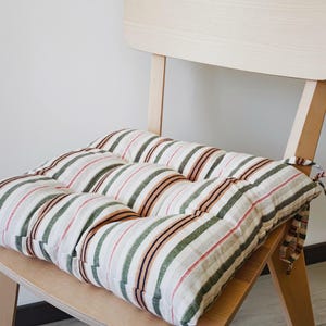 May include: A square seat cushion featuring a striped pattern in shades of olive green, rust orange, and dark blue on a white background. The cushion is tufted with visible stitching and includes fabric ties on the side for securing to a chair.