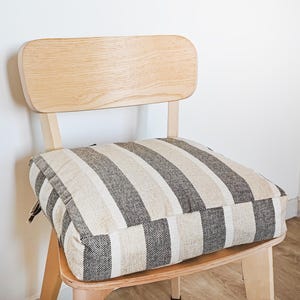 May include: A wooden chair with a light-colored finish, featuring a square cushion with vertical stripes in shades of beige, gray, and white. The chair back is a curved, rectangular shape. The chair is set against a white wall.