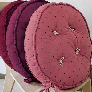 Puede incluir: Tres cojines de asiento redondos y capitoné en tonos rosa empolvado, ciruela intenso y burdeos oscuro. Cada cojín presenta un patrón de pequeños lunares negros, un borde ribeteado y tres botones centrales sujetos con lazos de tela clara.