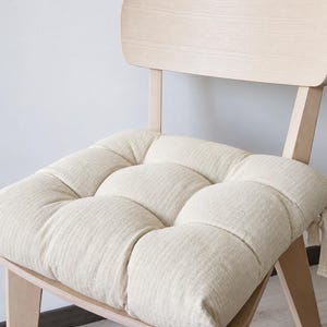 Könnte beinhalten: Ein quadratisches, cremeweißes Sitzkissen mit einem gesteppten Design, das aus neun einzelnen, gepolsterten Abschnitten besteht. Das Kissen ist aus strukturiertem Stoff gefertigt und verfügt über seitliche Stoffbänder zur Befestigung an einem hellen Holzstuhl.