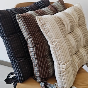 Puede incluir: Tres cojines cuadrados para silla con rayas verticales y detalles de cuadros. Los cojines son de color negro, marrón oscuro y crema, cada uno con lazos de tela a juego en las esquinas para sujetarlos al asiento, dispuestos en fila.
