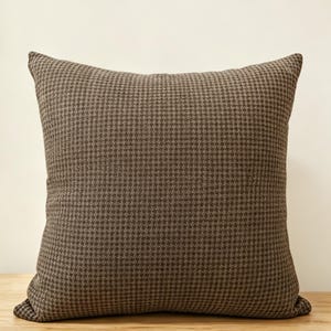 May include: Square decorative pillow with a brown and dark brown houndstooth pattern. The pillow is made of a textured fabric and sits on a wooden surface. The pillow has a classic, neutral design.