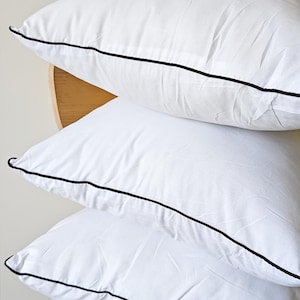 Puede incluir: Tres almohadas blancas apiladas, con ribete negro. Las almohadas parecen suaves y mullidas, sugiriendo un juego de cama cómodo. Imagen sencilla y limpia, ideal para un dormitorio.