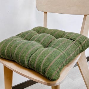 Puede incluir: Un cojín de asiento cuadrado con tela verde texturizada y rayas verticales marrones. El cojín está acolchado con nueve secciones distintas y se coloca sobre una silla de madera clara con respaldo curvo y cuatro patas.