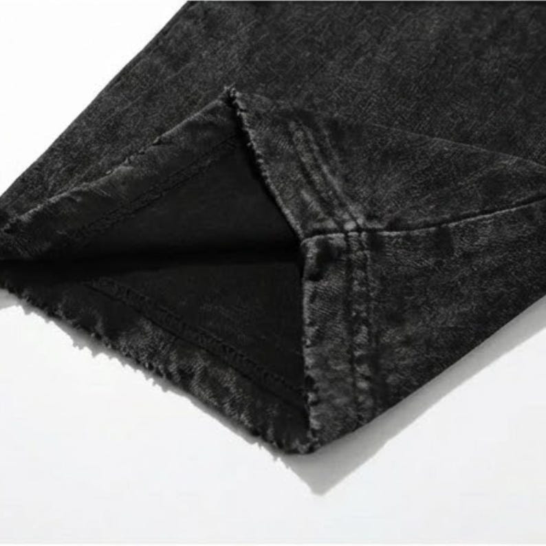 Puede incluir: Primer plano de unos vaqueros de mezclilla negros con un aspecto descolorido y desgastado. La tela tiene un aspecto texturizado y el dobladillo es visible. Los vaqueros est&aacute;n doblados, mostrando el forro interior y las costuras.