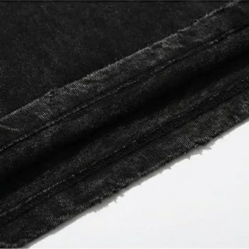 Puede incluir: Primer plano de tela vaquera negra, que muestra la textura y los detalles de las costuras. El denim tiene un aspecto descolorido, con costuras visibles y un borde sin rematar. La tela est&aacute; ligeramente arrugada, lo que sugiere que es parte de una prenda.