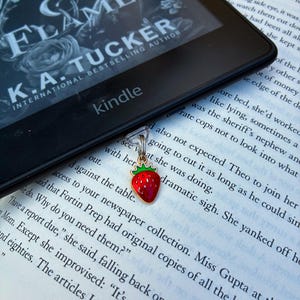 May include: A small strawberry-shaped charm with a red body, green leaves, and gold-tone metal edges, attached to a clear plastic clip. The charm rests on an open book page next to a black Kindle e-reader displaying the book title Fate of Wrath & Flame by K.A. Tucker.
