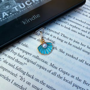 May include: A gold-toned metal bookmark charm shaped like a seashell with bright blue enamel inlay and a small white pearl at the center. The charm is attached to a clear plastic clip resting on an open book page near a black Kindle e-reader.