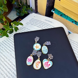 May include: A set of five metal charms with clear plastic clips, each featuring colorful enamel illustrations of marine life including a starfish, a seashell, an octopus, a shrimp, and a fish, arranged on a black electronic device.