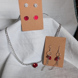 May include: A silver-toned chain necklace with a round red pendant, a pair of silver-toned stud earrings, a pair of red stud earrings, and a pair of dangling earrings featuring silver-toned chains and small red beads on brown display cards.