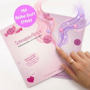 May include: A pink notepad with the text "Schnupperblock" and candy illustrations. The notepad has a "scratch and sniff" effect, with a finger pointing at the scented area. The text "Mhmm dieser Candyduft!" is also visible on the notepad.